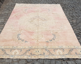 May include: A rectangular area rug featuring a soft pink background with a large, intricate central medallion in cream and beige. The rug is decorated with symmetrical floral motifs in blue and cream, and a wide border with blue and beige patterns.