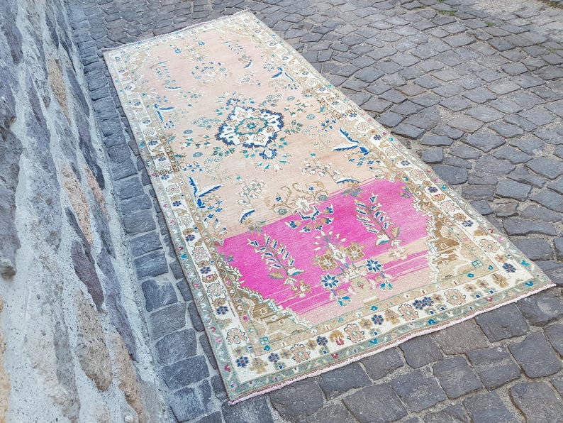 May include: A rectangular area rug featuring a faded peach background with a prominent bright pink section at one end. The rug displays intricate floral and geometric patterns in shades of blue, cream, and brown, finished with a decorative border.
