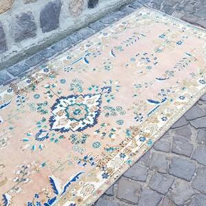May include: A rectangular area rug featuring a light peach background with a central medallion in navy blue, white, and teal. The rug is decorated with intricate floral and vine patterns in shades of blue, green, and tan, finished with a patterned border.