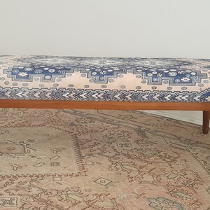May include: A wooden bench with a blue and pink patterned cushion. The bench has turned wooden legs and is sitting on a patterned rug.