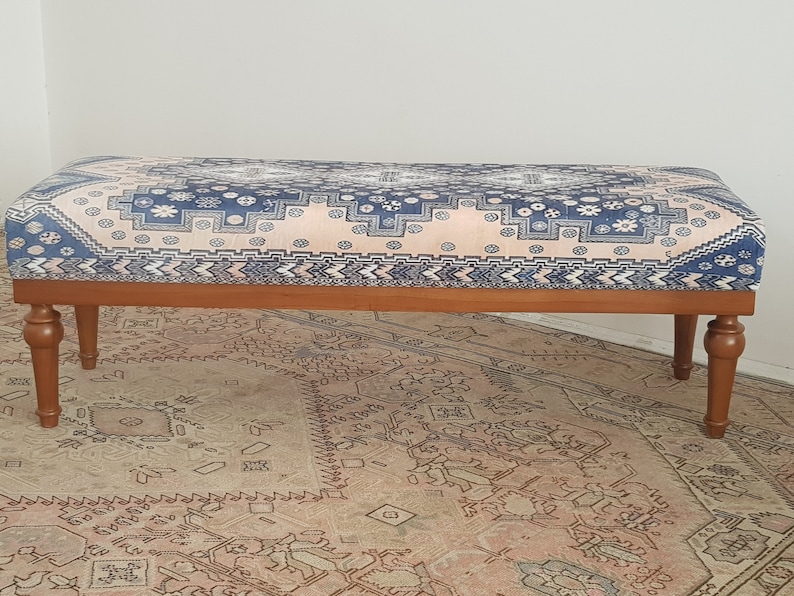 May include: A wooden bench with a blue and pink patterned cushion. The bench has turned wooden legs and is sitting on a patterned rug.