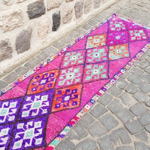 May include: A vibrant, rectangular runner rug with a pink background and geometric patterns in purple, orange, and teal. The rug is placed on a cobblestone surface next to a stone wall.