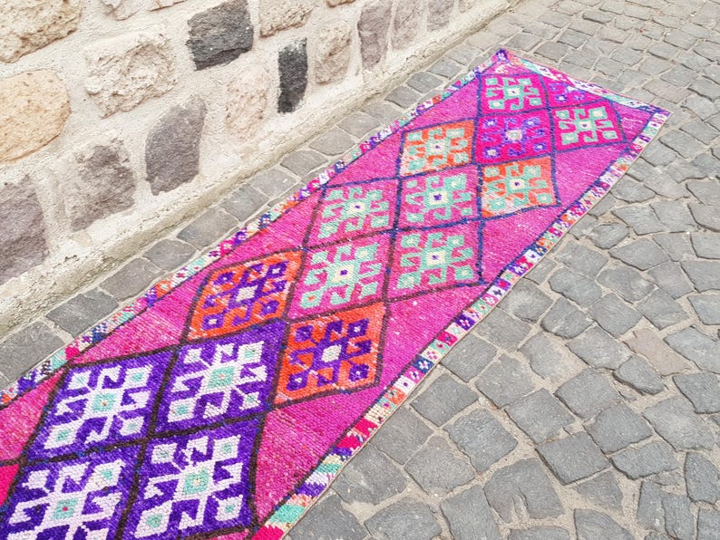 May include: A vibrant, rectangular runner rug with a pink background and geometric patterns in purple, orange, and teal. The rug is placed on a cobblestone surface next to a stone wall.