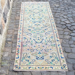 Op de afbeelding: Een rechthoekig vintage tapijt met een lichtgroene en beige achtergrond. Het tapijt heeft een gedetailleerd bloemenpatroon in blauw, groen en goud. Het tapijt ligt op een kasseien oppervlak.