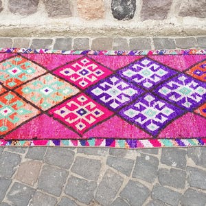 May include: A vibrant, patterned runner rug with a repeating diamond design. The rug features a bright pink background with geometric shapes in orange, purple, and teal. The rug is likely made of woven material.