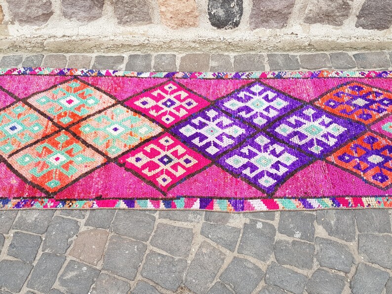 May include: A vibrant, patterned runner rug with a repeating diamond design. The rug features a bright pink background with geometric shapes in orange, purple, and teal. The rug is likely made of woven material.