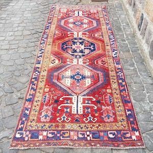 Pode incluir: Um tapete retangular de estilo vintage com um padrão geométrico central em vermelho, azul marinho e creme. O tapete apresenta intrincados desenhos de borda e uma aparência desgastada, sugerindo que é uma antiguidade ou uma reprodução.