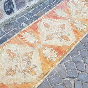 May include: A long, rectangular runner rug featuring a vibrant orange background with repeating cream-coloured diamond-shaped medallions. The rug displays intricate floral and geometric patterns in shades of orange, cream, and light green throughout.