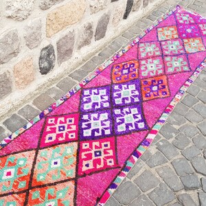 May include: A vibrant, rectangular rug with a repeating diamond pattern in shades of pink, purple, orange, and teal. The rug is placed on a stone surface, showcasing its intricate design and color palette.