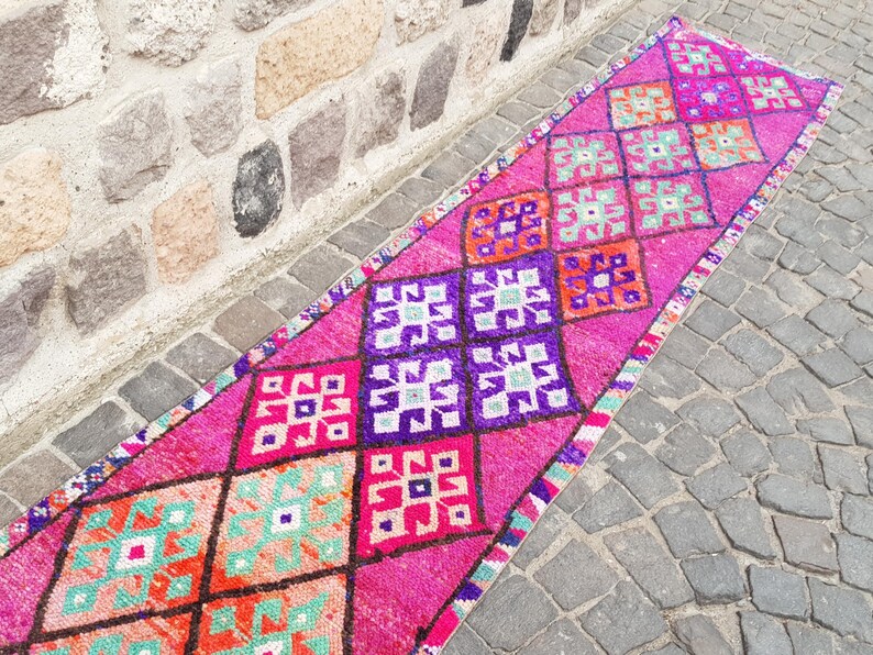 May include: A vibrant, rectangular rug with a repeating diamond pattern in shades of pink, purple, orange, and teal. The rug is placed on a stone surface, showcasing its intricate design and color palette.