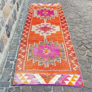 May include: A long rectangular runner rug featuring a vibrant orange background with three large, symmetrical geometric medallions in shades of hot pink, brown, and cream. The border displays a repeating yellow zigzag pattern on a red base.