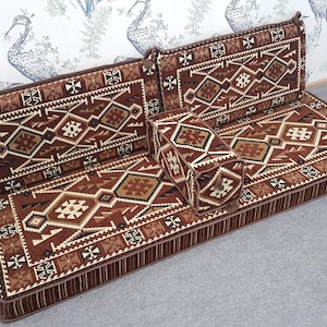 May include: A brown and beige patterned floor cushion set with two backrests and a bolster. The cushions feature a geometric design with a variety of shapes and colors. The set is placed on a gray surface.