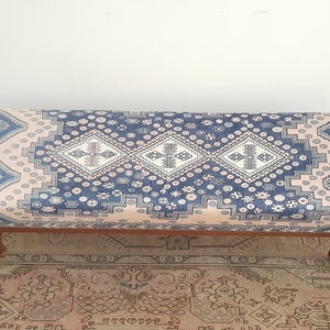 May include: A long, rectangular ottoman with a pink and blue patterned fabric top. The ottoman has four wooden legs.