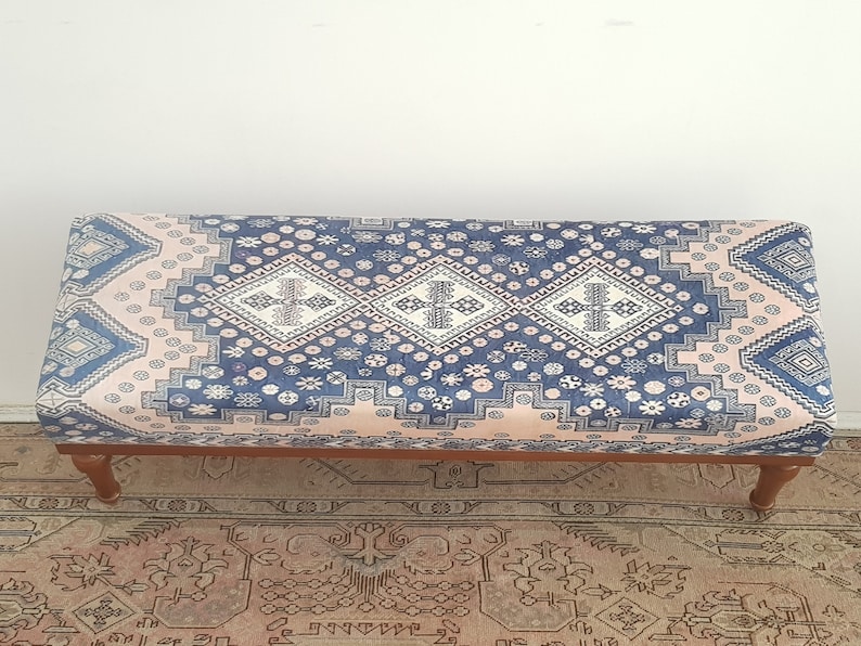 May include: A long, rectangular ottoman with a pink and blue patterned fabric top. The ottoman has four wooden legs.
