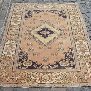 Puede incluir: Una alfombra rectangular que presenta un medallón central en forma de diamante en azul oscuro y crema sobre un campo de color melocotón. La alfombra está decorada con motivos florales y geométricos repetitivos, enmarcada por un borde ancho.