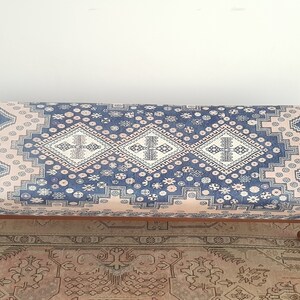 May include: A blue and pink patterned ottoman with wooden legs. The ottoman has a traditional design with geometric shapes and floral motifs.