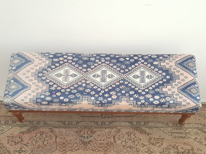 May include: A blue and pink patterned ottoman with wooden legs. The ottoman has a traditional design with geometric shapes and floral motifs.
