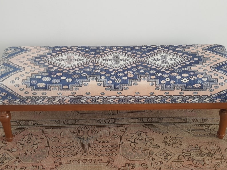 May include: A wooden bench with a blue and pink patterned cushion. The cushion has a geometric design with white accents. The bench is sitting on a rug with a similar pattern.