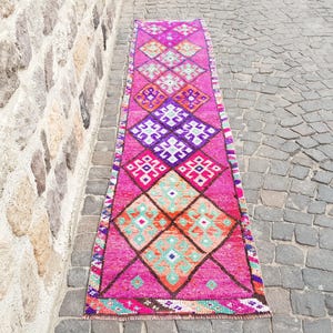 May include: A vibrant, rectangular runner rug with a repeating diamond pattern in shades of pink, purple, orange, and turquoise. The rug is placed on a cobblestone surface, showcasing its intricate design and color palette.