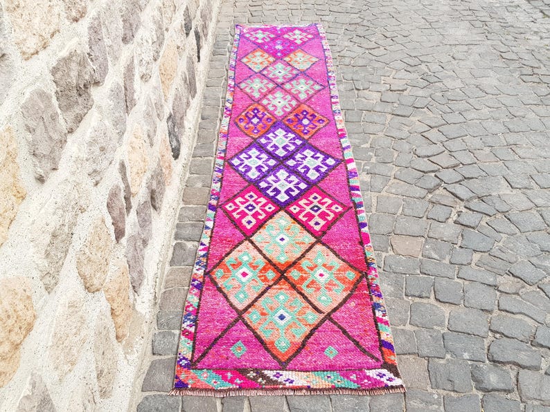 May include: A vibrant, rectangular runner rug with a repeating diamond pattern in shades of pink, purple, orange, and turquoise. The rug is placed on a cobblestone surface, showcasing its intricate design and color palette.