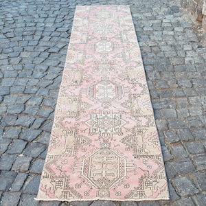 May include: A long, rectangular runner rug featuring a soft pink background with intricate geometric patterns in shades of beige, cream, and dark brown. The design includes repeating central medallions and detailed border motifs throughout the surface.