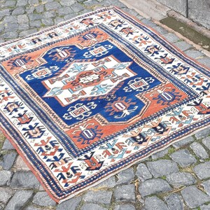 May include: A rectangular area rug featuring a central diamond medallion in white, orange, and blue. The rug displays a geometric pattern with intricate borders in shades of navy blue, cream, and burnt orange, finished with short fringe on the edges.