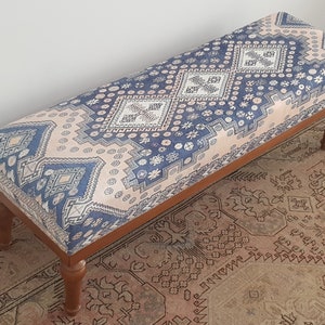 May include: A wooden bench with a blue and pink patterned cushion. The cushion has a geometric design with diamonds and flowers. The bench is sitting on a rug with a similar pattern.