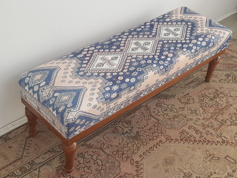May include: A wooden bench with a blue and pink patterned cushion. The cushion has a geometric design with diamonds and flowers. The bench is sitting on a rug with a similar pattern.