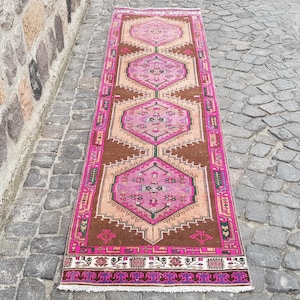 May include: A long, rectangular rug with a geometric pattern in shades of pink, brown, and beige. The rug features a central design with diamond-shaped motifs and a border with intricate detailing. The rug is placed on a cobblestone surface.