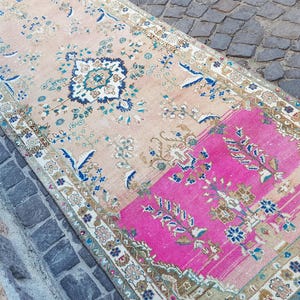May include: A rectangular area rug featuring a faded peach background with a prominent section of vibrant hot pink. The rug is decorated with intricate floral and geometric patterns in shades of navy blue, cream, and muted brown throughout.
