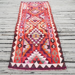 May include: A rectangular runner rug featuring a vibrant geometric pattern in shades of red, orange, purple, pink, and white. The design consists of repeating diamond shapes and triangular motifs arranged in a symmetrical, linear composition.