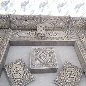 May include: A gray sectional sofa with matching cushions and a small square ottoman. The sofa and cushions feature a geometric pattern in shades of gray, black, and white. The set is arranged on a gray surface, with a patterned wallpaper in the background.