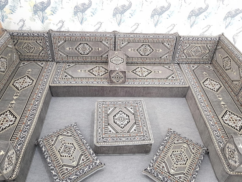 May include: A gray sectional sofa with matching cushions and a small square ottoman. The sofa and cushions feature a geometric pattern in shades of gray, black, and white. The set is arranged on a gray surface, with a patterned wallpaper in the background.