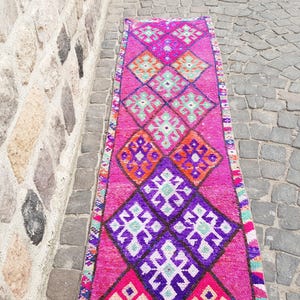 May include: A vibrant, rectangular rug with a repeating diamond pattern in shades of pink, purple, orange, and teal. The rug is placed on a stone surface next to a stone wall. The design features geometric shapes and a border.