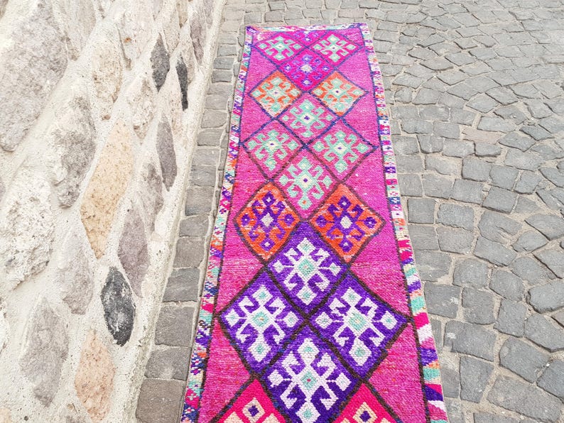 May include: A vibrant, rectangular rug with a repeating diamond pattern in shades of pink, purple, orange, and teal. The rug is placed on a stone surface next to a stone wall. The design features geometric shapes and a border.