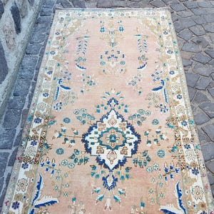 May include: A rectangular area rug featuring a soft peach background with a central dark blue and white floral medallion. The rug is decorated with symmetrical botanical patterns in shades of blue, green, and beige, framed by a detailed border.