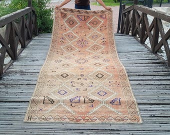 May include: A long, rectangular runner rug featuring a faded peach and beige background with a repeating pattern of large diamonds. The design includes smaller geometric motifs in shades of dark brown, blue, and purple, with a zigzag border.