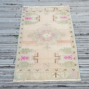 May include: A rectangular area rug featuring a light beige base with a central geometric medallion and corner motifs in muted sage green, soft brown, and vibrant fuchsia pink. The rug has a decorative border and short fringe along the edges.