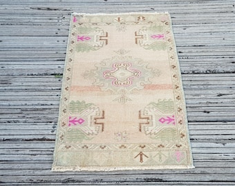 May include: A rectangular area rug featuring a light beige base with a central geometric medallion and corner motifs in muted sage green, soft brown, and vibrant fuchsia pink. The rug has a decorative border and short fringe along the edges.