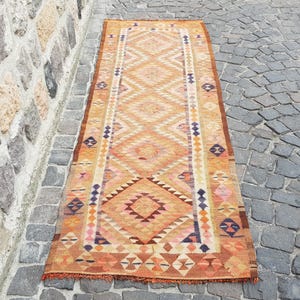 May include: A long, rectangular runner rug featuring a repeating diamond pattern in shades of orange, yellow, and brown. The rug includes small geometric accents in blue, pink, and cream, with a fringed edge at the bottom and a border design.