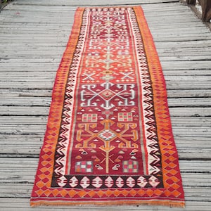 May include: A long, rectangular runner rug featuring a vibrant red base color with intricate geometric patterns in white, orange, and dark brown. The design includes repeating symmetrical motifs, checkered squares, and a decorative border.