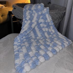 Op de afbeelding: Een zachte, getextureerde babydeken in een blauw-wit ruitpatroon. De deken is gemaakt van een zacht materiaal, met een pom-pom-achtige textuur. De deken is opgevouwen en toont het patroon en de textuur.