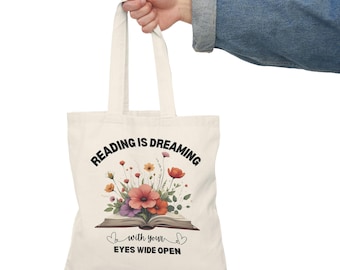 May include: Cream-colored tote bag with the text "READING IS DREAMING" above an illustration of an open book with colorful flowers. The text "...with your EYES WIDE OPEN" is below the book. The bag has two handles.