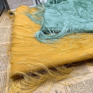 Puede incluir: Dos piezas de tela con flecos. Una pieza es de color amarillo mostaza y la otra es de color verde claro.