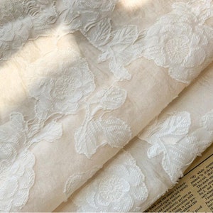 May include: A close-up of a white fabric with a delicate floral pattern. The fabric is sheer and has a soft, flowing texture.