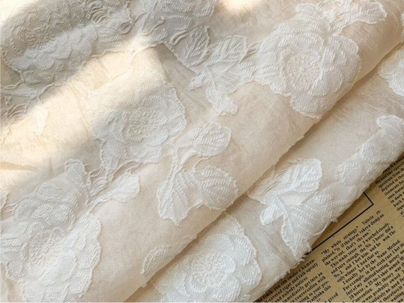 May include: A close-up of a white fabric with a delicate floral pattern. The fabric is sheer and has a soft, flowing texture.