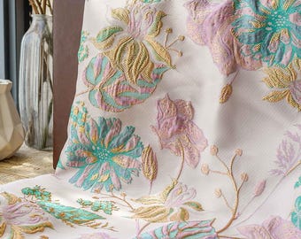 May include: Cream-colored fabric with a floral pattern in shades of teal, lavender, and gold. The design features large, stylized flowers and leaves with intricate detailing. The fabric appears to be a decorative textile.