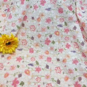 May include: White eyelet fabric with a floral pattern in shades of pink, orange, and purple. The fabric has a delicate, airy appearance with small, evenly spaced holes. A large yellow flower is in the lower left corner.