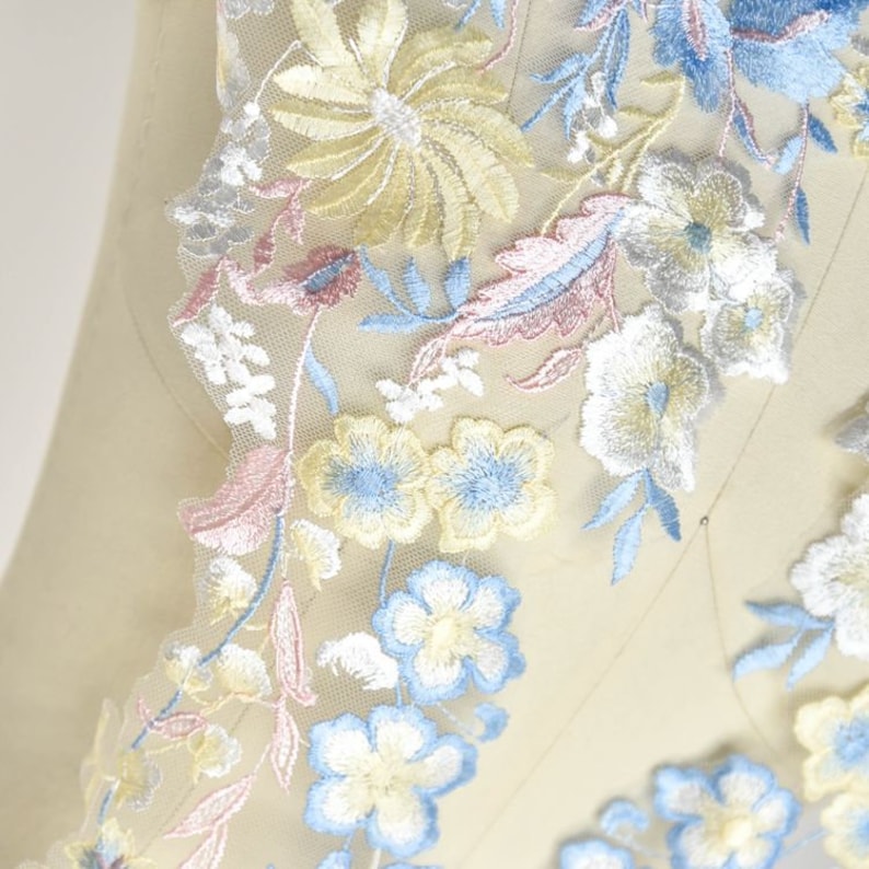 May include: A close-up of a delicate floral lace trim with a scalloped edge. The lace features a variety of pastel flowers, including yellow, blue, and pink, embroidered on a sheer mesh fabric.