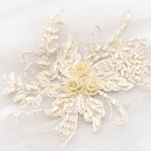 May include: A delicate white lace applique with a cluster of off-white pearl-like flowers in the center.